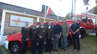 A group of men in uniforms and suits stand in front of a red fire truck with a banner behind them.