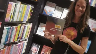 A woman in a bookstore holding a book titled 'Leben im Rhythmus der Natur', smiling at the camera.