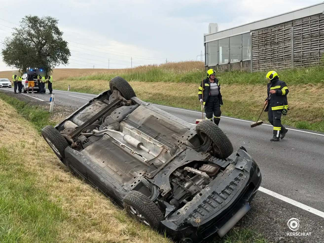 Ein umgekipptes Auto am Straßenrand mit zwei Feuerwehrleuten in der Nähe.