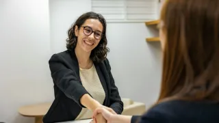 A smiling woman in a black blazer shakes hands with another woman in a professional setting.