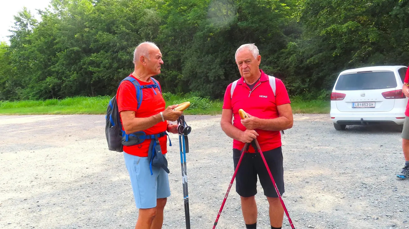 Zwei ältere Männer, beide in roten Shirts und mit Wanderstöcken, stehen auf einem Kiesweg. Sie halten beide ein Sandwich in den Händen. Der Mann auf der linken Seite hat einen Rucksack an.