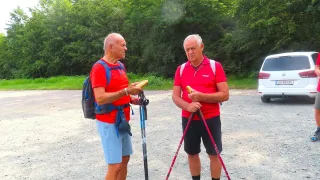 Two older men, both wearing red shirts and holding hiking poles, are standing on a gravel path. They are both holding a sandwich in their hands. The man on the left has a backpack on.