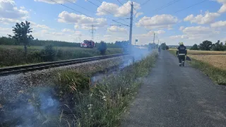 Ein Feuerwehrmann geht neben einer Bahnstrecke, wo ein kleiner Brand im Gras brennt. Ein roter Feuerwehrwagen ist in der Nähe geparkt.