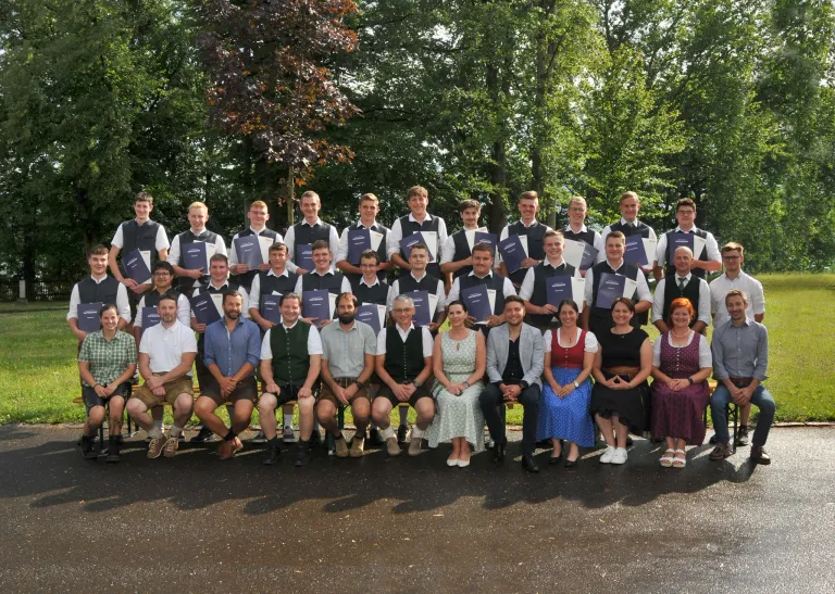 A group of people, both men and women, dressed in traditional attire, are posing for a photo in an outdoor setting with trees in the background. They are holding certificates and smiling for the camera.