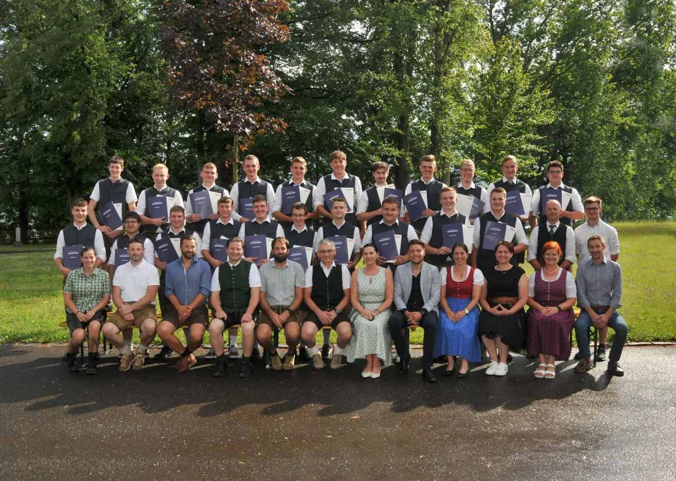 A group of people, both men and women, dressed in traditional attire, are posing for a photo in an outdoor setting with trees in the background. They are holding certificates and smiling for the camera.