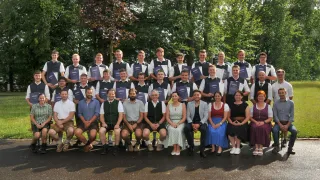 A group of people, both men and women, dressed in traditional attire, are posing for a photo in an outdoor setting with trees in the background. They are holding certificates and smiling for the camera.