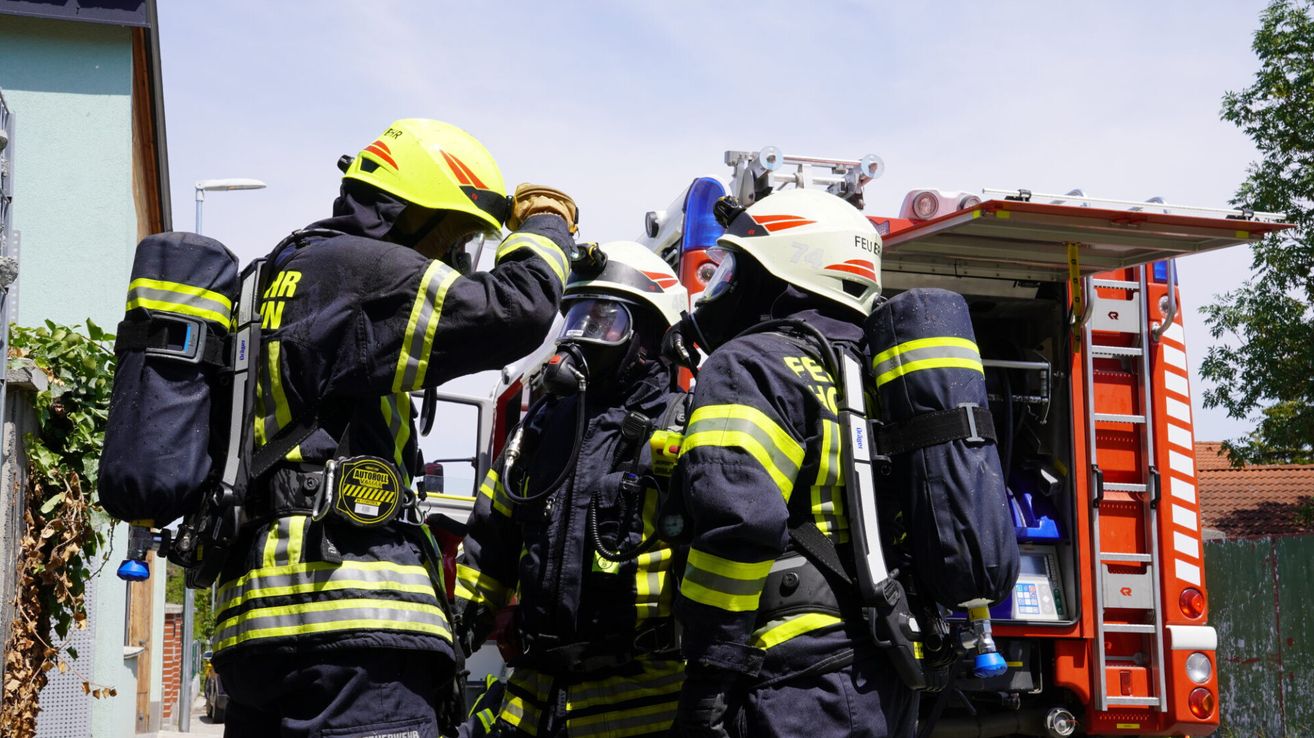 A group of firefighters in full gear, one adjusting a helmet, another with a mask, and a third with an oxygen tank. They are standing in front of a fire truck.