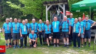 A group of older men in blue shirts and colorful ties pose for a photo in a park with a wooden structure and trees in the background.