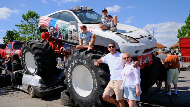 A family stands in front of a monster truck with a wrecked car on top. Two boys sit on the car, while a man and woman stand beside the truck.