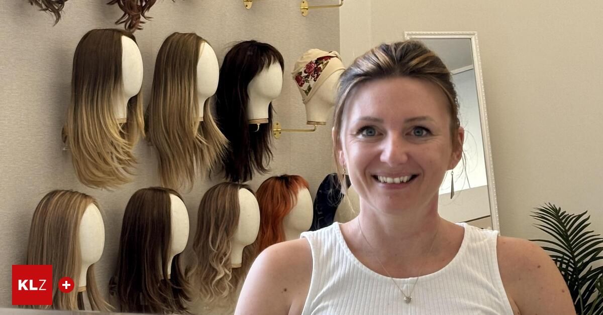 A woman stands in front of a wall with various wigs and mannequin heads, smiling for a photo.