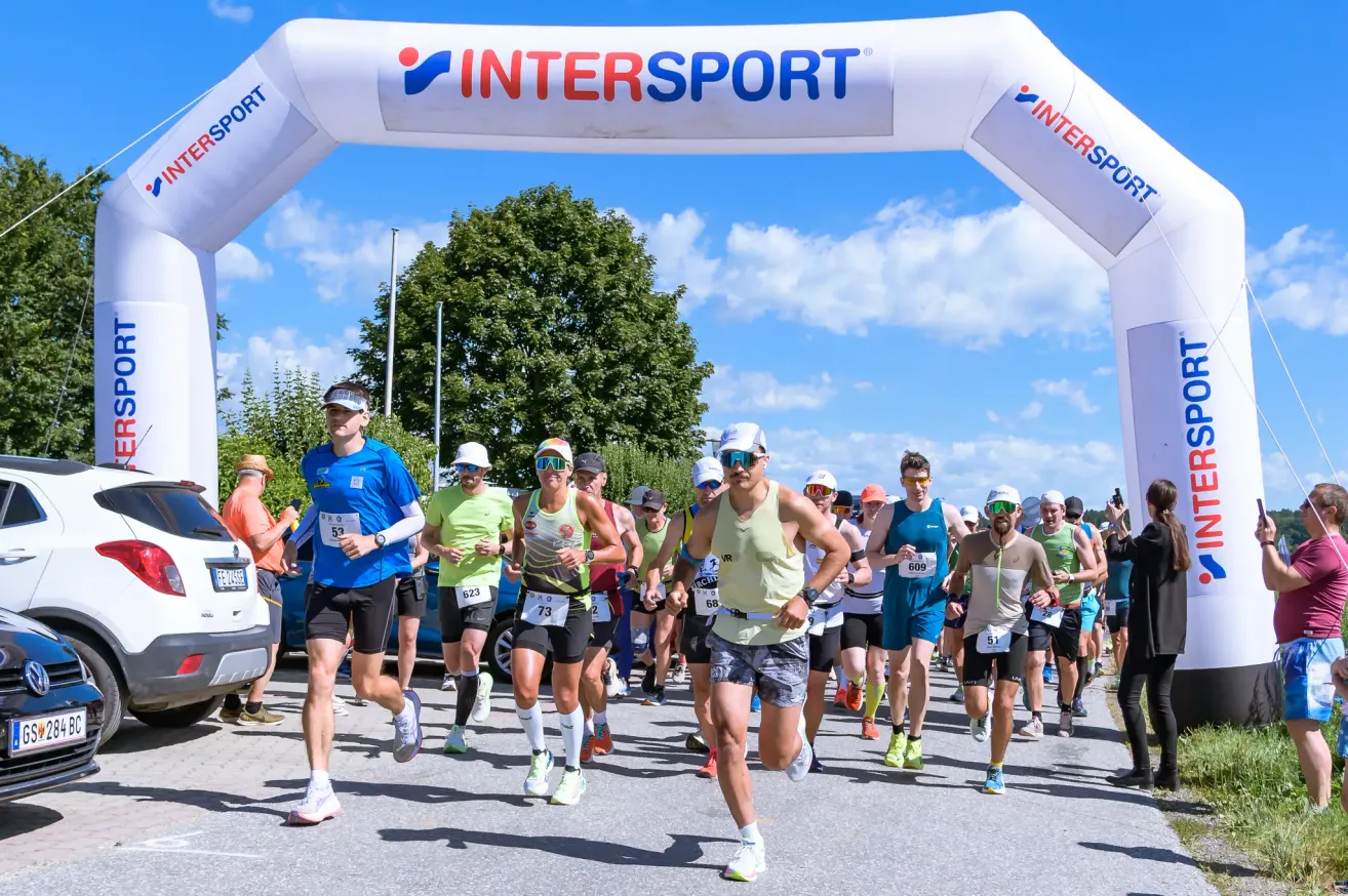 Eine Gruppe von Läufern mit nummerierten Startnummern nimmt an einem Marathon teil, unter einem Bogen mit dem Wort Intersport.