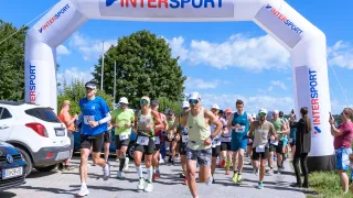 Eine Gruppe von Läufern mit nummerierten Startnummern nimmt an einem Marathon teil, unter einem Bogen mit dem Wort Intersport.