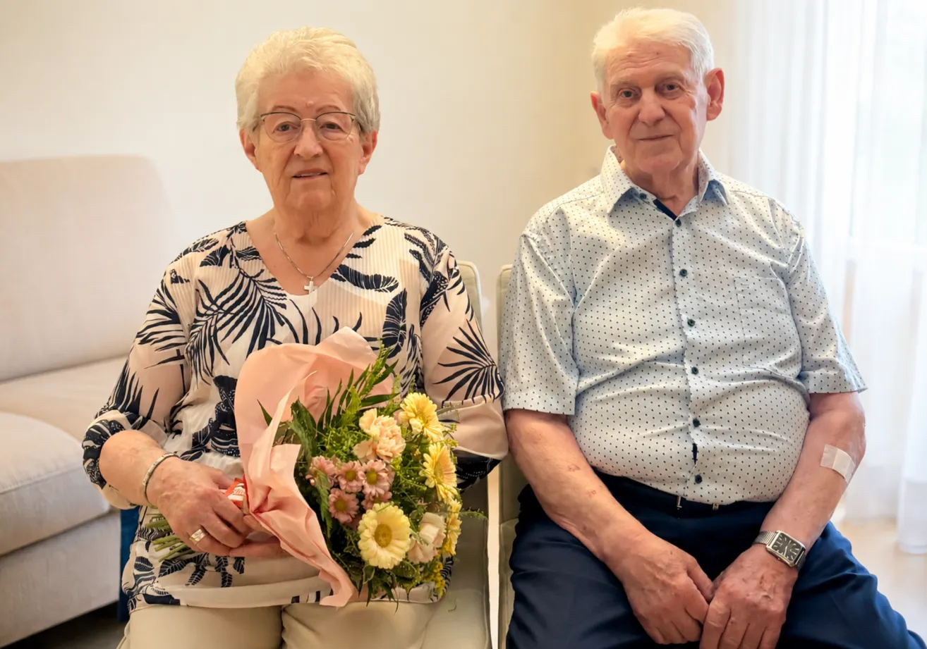 Ein älteres Paar sitzt nebeneinander, die Frau hält einen Blumenstrauß in der Hand, beide lächeln in die Kamera.