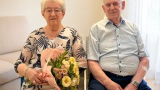 Ein älteres Paar sitzt nebeneinander, die Frau hält einen Blumenstrauß in der Hand, beide lächeln in die Kamera.