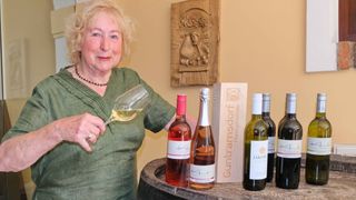 An older woman in a green dress is holding a glass of wine and standing next to a table with several wine bottles and a wooden box.