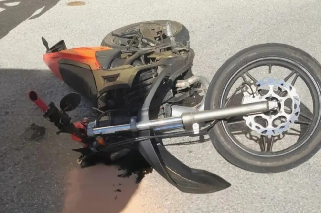 A motorcycle lies on its side on the road, with its front wheel detached and the engine exposed. The motorcycle's body is orange and black, and it has a silver handlebar.