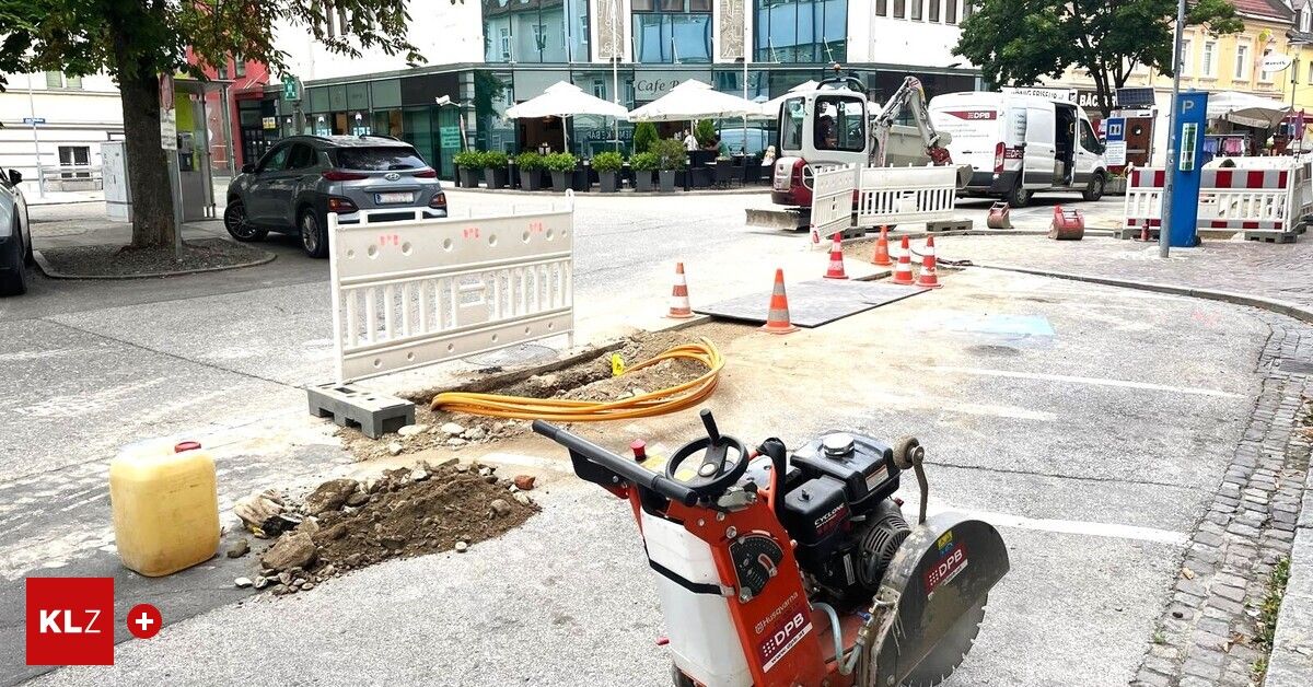 A construction site with a sawing machine, traffic cones, and a parked car on a street with a building and parked vehicles in the background.