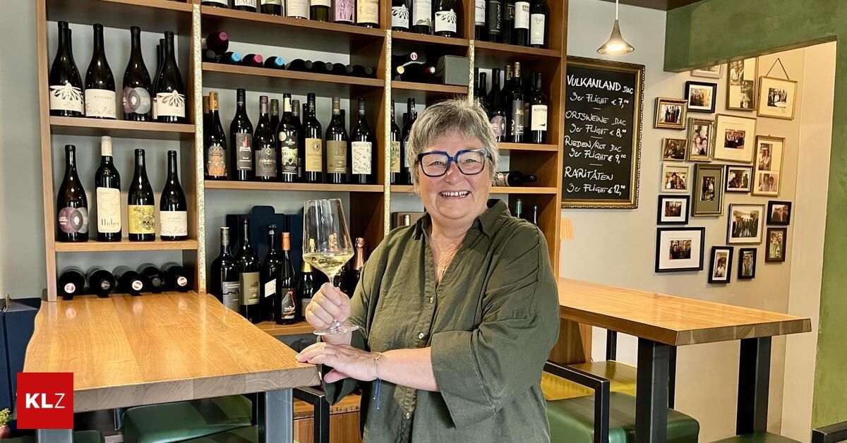 A woman in a green shirt holds a wine glass in a wine shop with shelves full of wine bottles in the background.