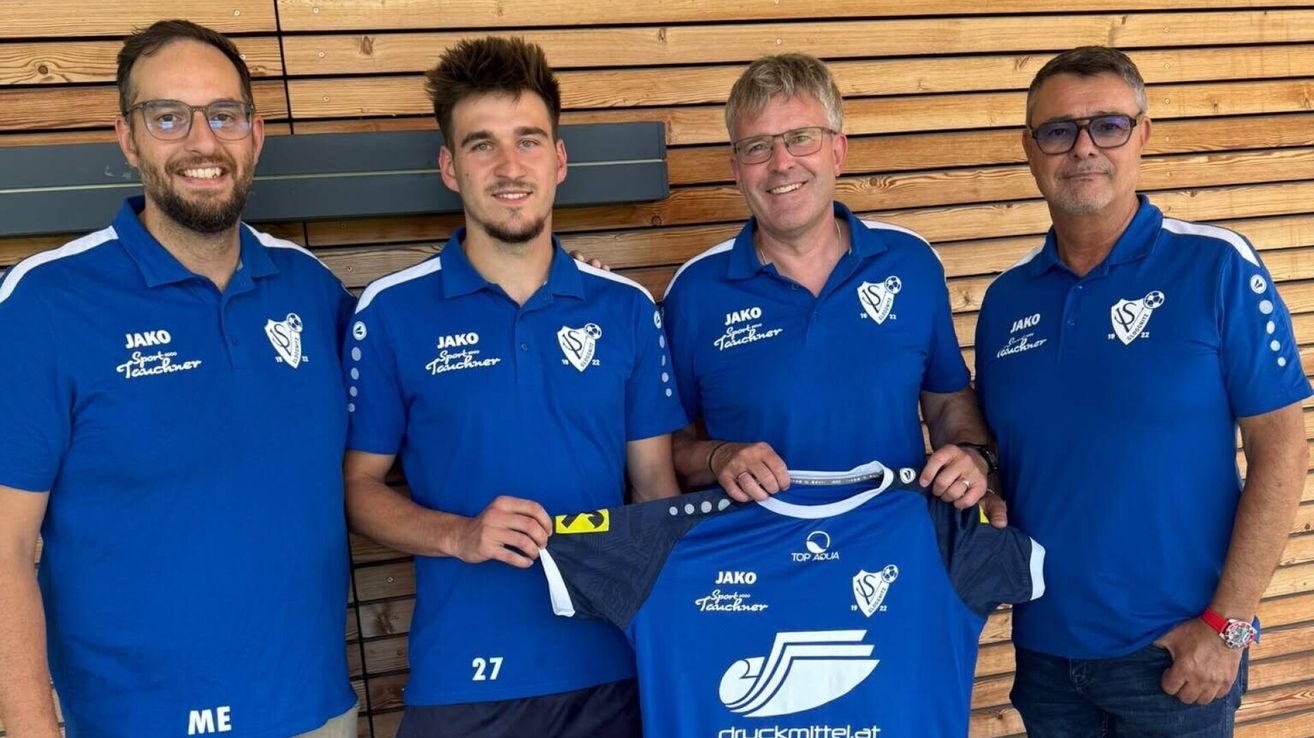 Four men in blue sports jerseys with white trims and logos on the chest. The man in the middle holds up a blue jersey with white text and logos. The wooden wall behind them has a gray frame.