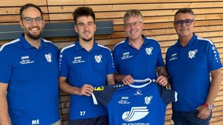 Four men in blue sports jerseys with white trims and logos on the chest. The man in the middle holds up a blue jersey with white text and logos. The wooden wall behind them has a gray frame.