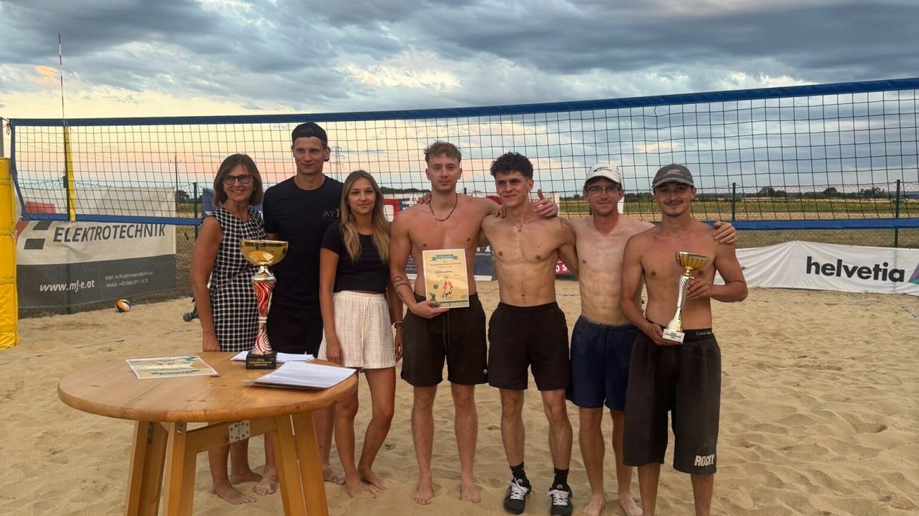 Eine Gruppe junger Männer und Frauen posiert für ein Foto am Strand mit einem Volleyballnetz im Hintergrund. Sie halten Pokale und Urkunden.
