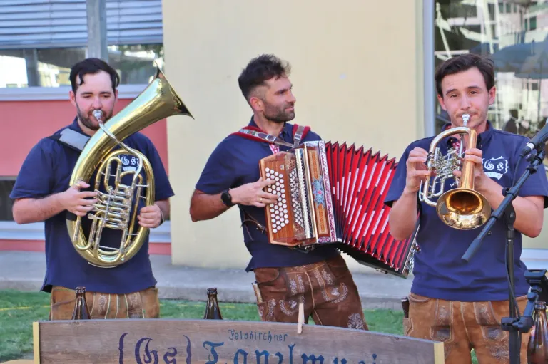 Three men in traditional attire play brass instruments in front of a building. The middle man plays an accordion, while the others play trumpets.