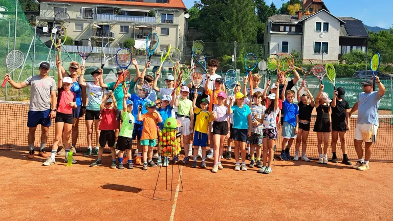 Eine Gruppe von Kindern und Erwachsenen steht auf einem Tennisplatz, hält Tennisschläger und posiert für ein Foto. Der Tennisplatz hat ein Netz in der Mitte, und ein Ballhalter mit Tennisbällen steht vor der Gruppe.