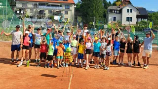 A group of children and adults are standing on a tennis court, holding tennis rackets, and posing for a photo. The tennis court has a net in the middle, and a ball holder with tennis balls is placed in front of the group.