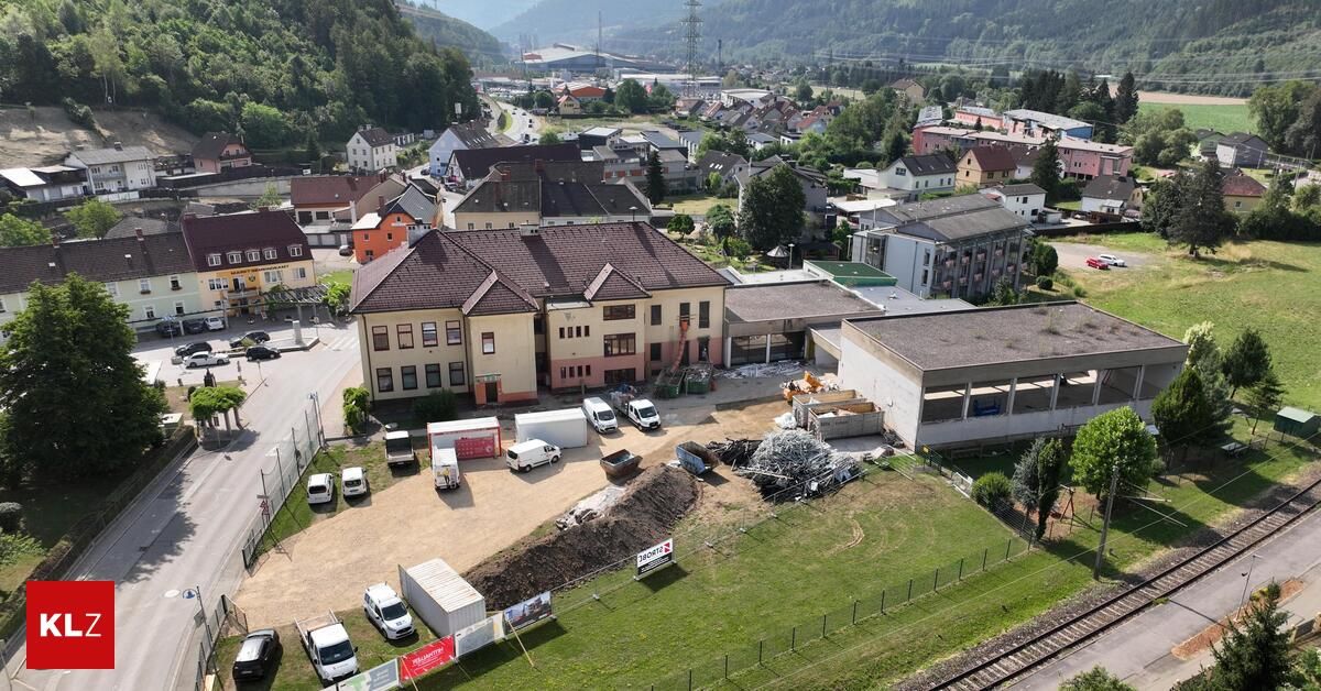 Aerial view of a town with a construction site in front of a large building, surrounded by various vehicles and greenery.
