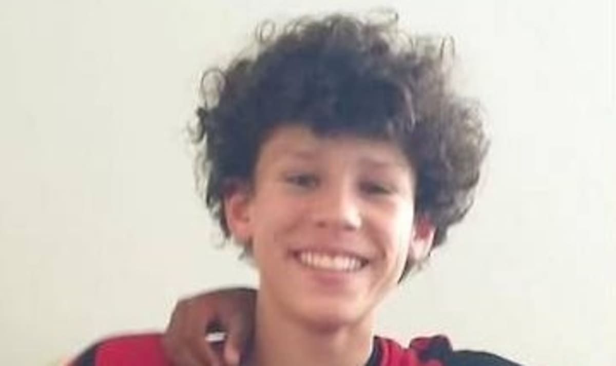 A young boy with curly hair is smiling and posing for a photo. He is wearing a red and black shirt.