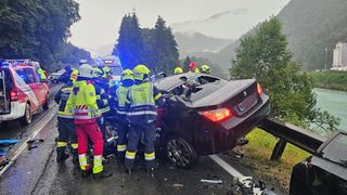 Emergency responders in yellow and red uniforms attend to a damaged black BMW on a road with a mountainous backdrop.
