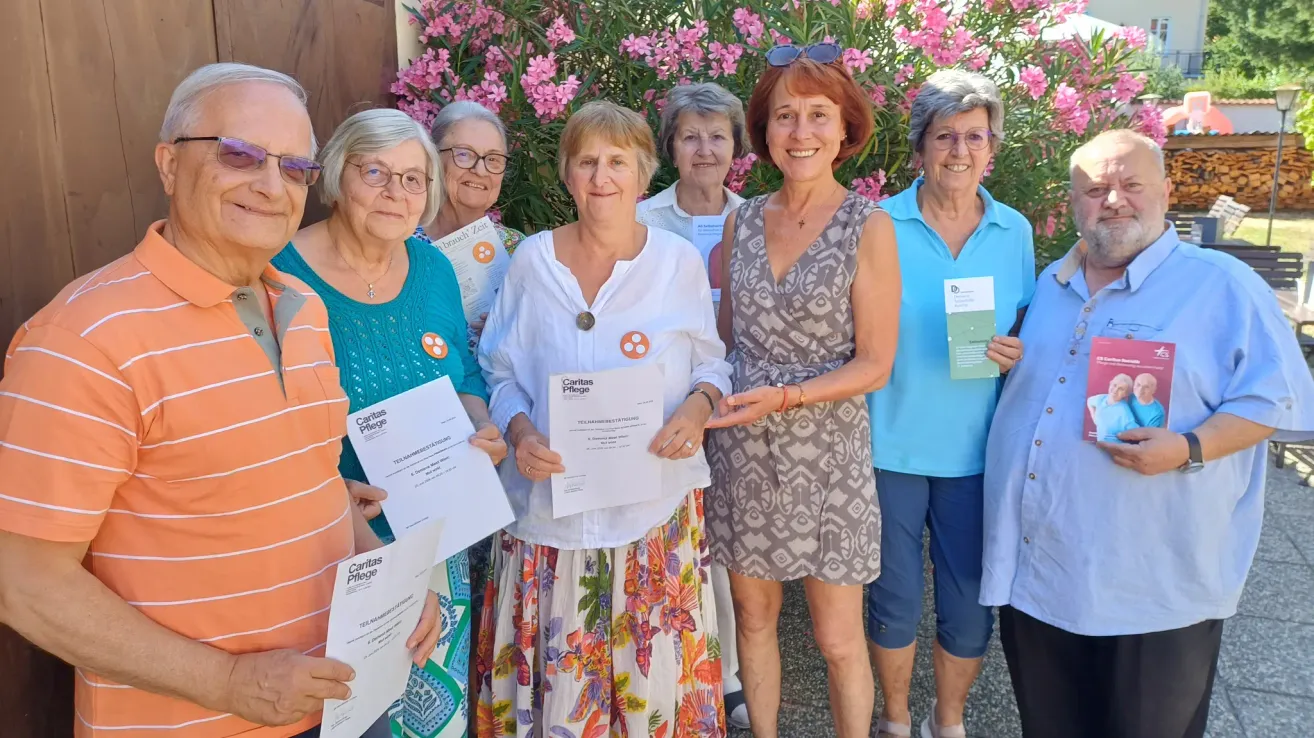A group of elderly women, all smiling, stand together holding certificates. They are in front of a bush with pink flowers.