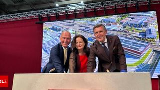 Drei Menschen in formeller Kleidung stehen hinter einem Podium und lächeln für ein Foto, wobei ein großer Bildschirm im Hintergrund eine Stadtlandschaft zeigt.