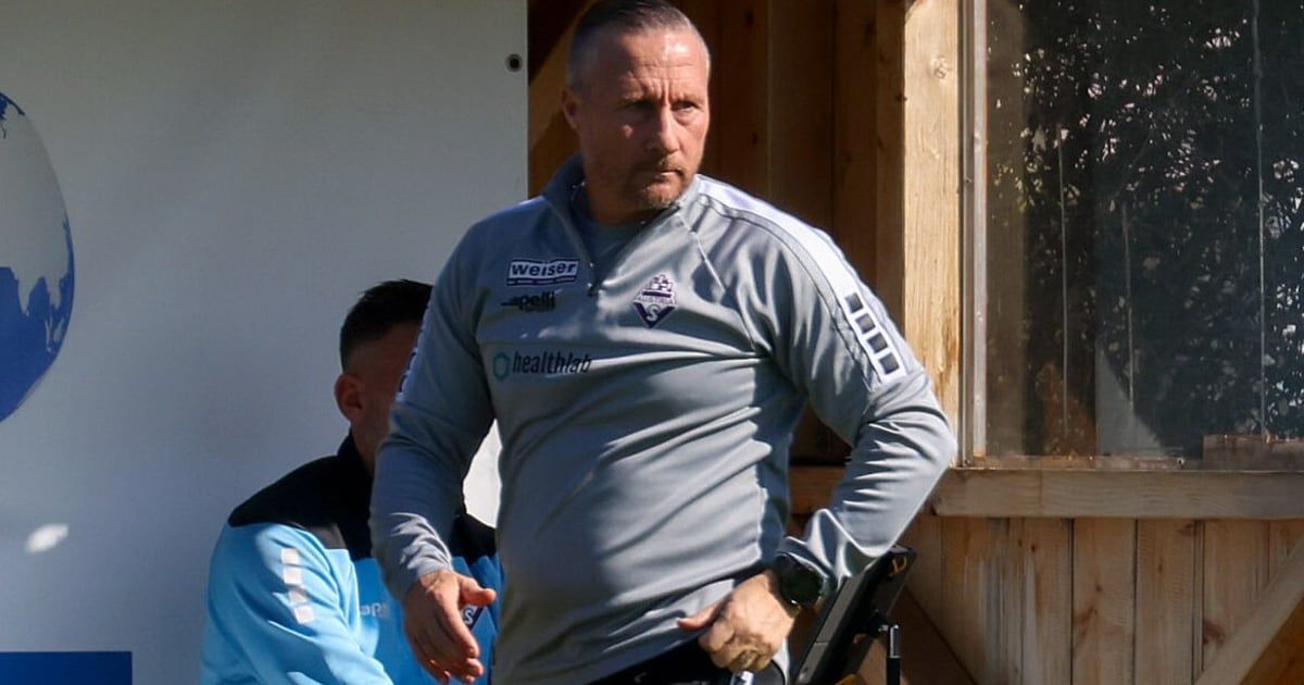 A man in a gray sports jacket with white stripes and logos walks outside, looking serious. Behind him, another man in a blue jacket stands near a wooden wall.