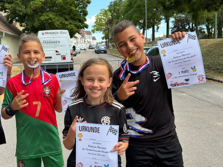 Three children stand together, smiling, holding certificates and medals. The boy on the right wears a medal around his neck. The girl in the middle holds a certificate with a car race design. The boy on the left wears a red jersey.