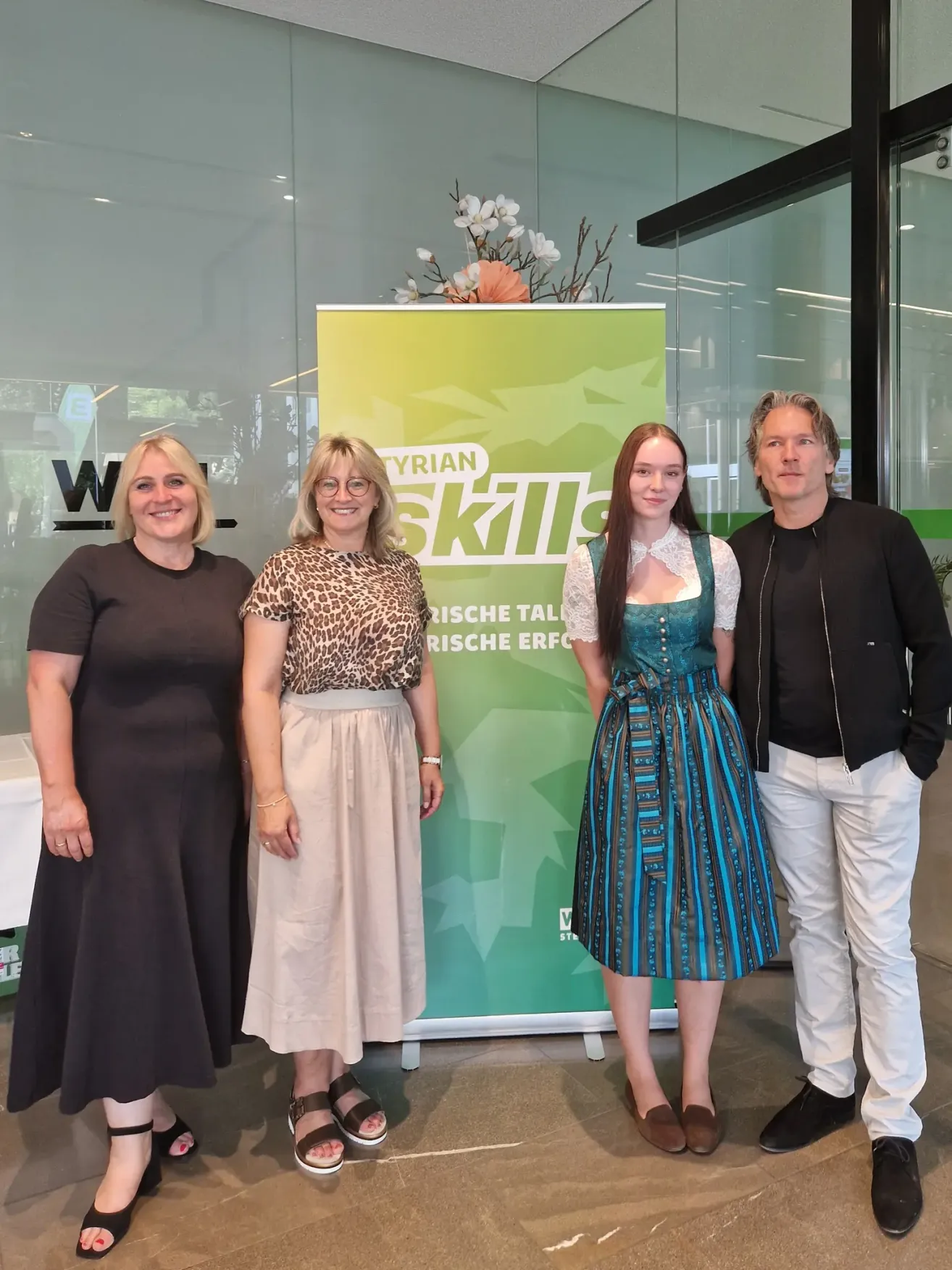 Vier Erwachsene, zwei Frauen und zwei Männer, stehen vor einem grünen Banner mit dem Wort 'Skill' darauf. Die Frau links trägt ein schwarzes Kleid, die Frau in der Mitte trägt ein Leopardenmuster-Oberteil und einen beigen Rock, die Frau rechts trägt ein blau-weißes Kleid und der Mann rechts trägt eine schwarze Jacke.