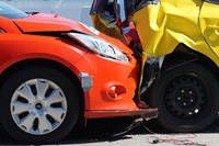Nahaufnahme eines beschädigten orangefarbenen Autos mit verbogenem Stoßfänger und einem gelben Auto mit beschädigter Vorderseite, beide auf einer Straße geparkt.
