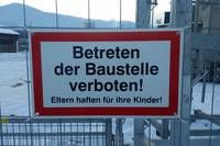Ein rotes und weißes Schild an einem Zaun steht 'Betreten der Baustelle verboten!' mit einem Hinweis 'Eltern haften für ihre Kinder!' in einer verschneiten Landschaft.