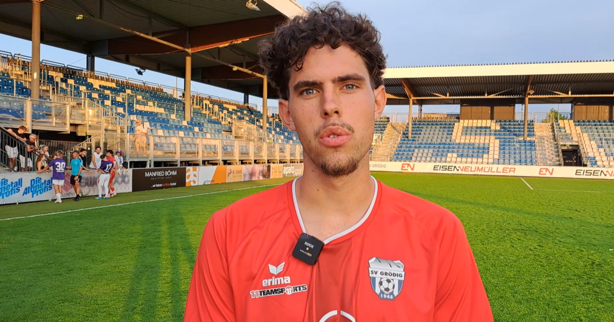 A soccer player with curly hair and a red jersey stands in an empty stadium with a green field and blue seats.