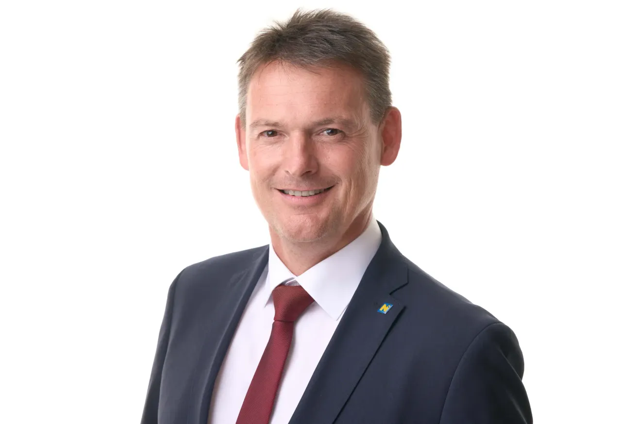 Ein Mann in Anzug und Krawatte, lächelt mit einem Abzeichen auf seiner Jacke.
