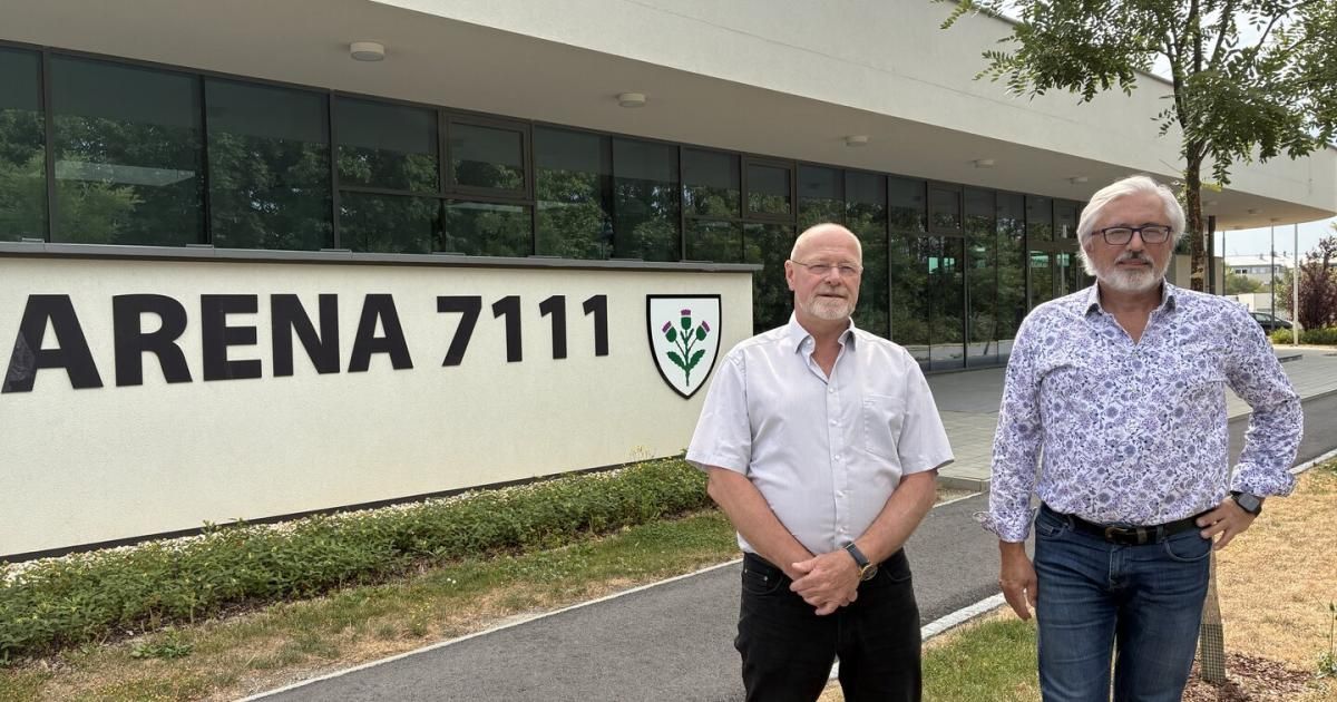 Zwei Männer stehen vor einem Gebäude mit einem Schild, auf dem 'ENA 7111' und ein Logo mit einem Distel stehen. Der Mann links trägt eine Brille und eine Uhr.