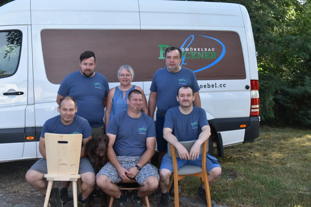 Five men and a woman in matching blue shirts pose in front of a van with the text 'ZMOEBELBAU' and 'oebel.cc' on it. A brown dog sits between two of the men.