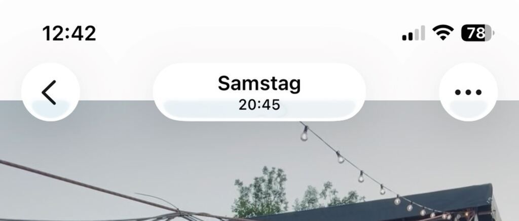 A screenshot of a phone displaying the day and time, with a string of lights hanging above a building in the background.