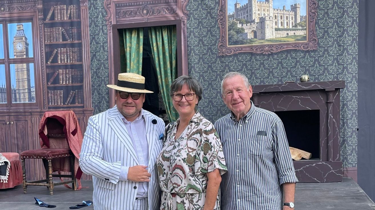 Drei Personen stehen vor einer Wand mit einem Schlossgemälde. Der Mann links trägt einen Strohhut und eine Sonnenbrille. Die Frau in der Mitte trägt eine Brille und ein Blumenhemd. Der Mann rechts trägt ein gestreiftes Hemd.