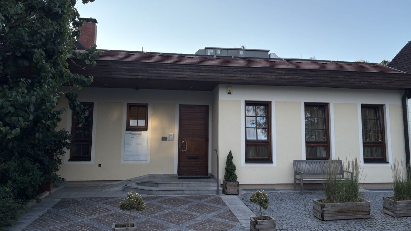A beige house with a brown door and three windows, surrounded by a brick pathway and small potted plants.