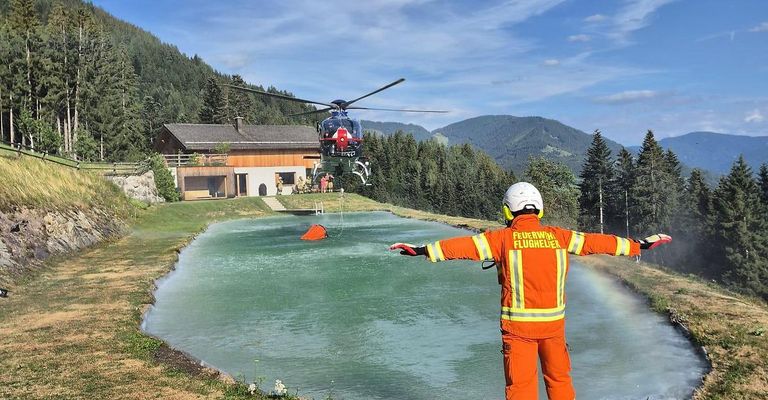 A firefighter in an orange uniform directs a helicopter to drop water into a pond. The helicopter is above a house with people on the porch.