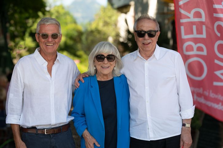 Drei Menschen, zwei Männer und eine ältere Frau, lächeln und posieren für ein Foto vor einem roten Banner.