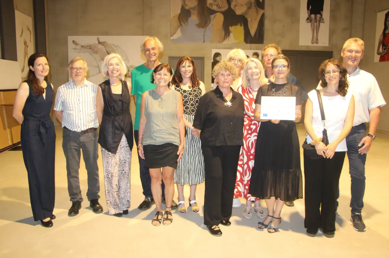 Eine Gruppe von Menschen steht in einem Raum, lächelt und posiert für ein Foto. Eine Frau hält ein Zertifikat.