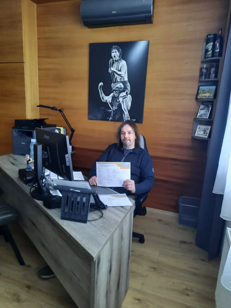 Ein Mann sitzt an einem Schreibtisch in einem Büro, hält ein Stück Papier in der Hand und lächelt. Hinter ihm ist eine Wand mit einem großen schwarz-weißen Poster.