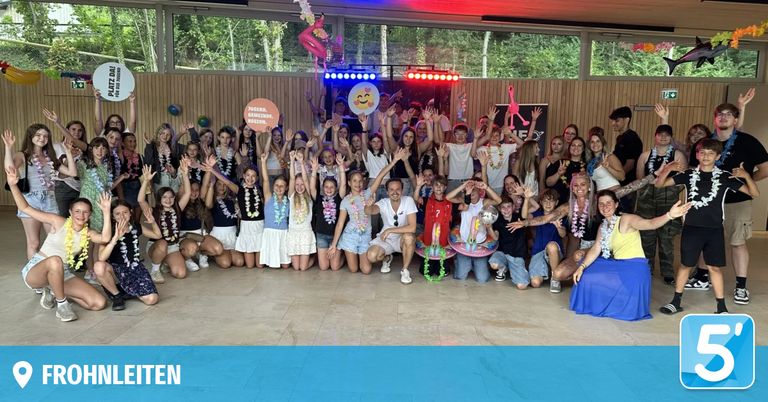 A group of people, possibly a team, are posing for a photo with smiles and raised hands. They are wearing garlands and some are holding balloons. The setting is indoors with a wooden wall and a disco light.